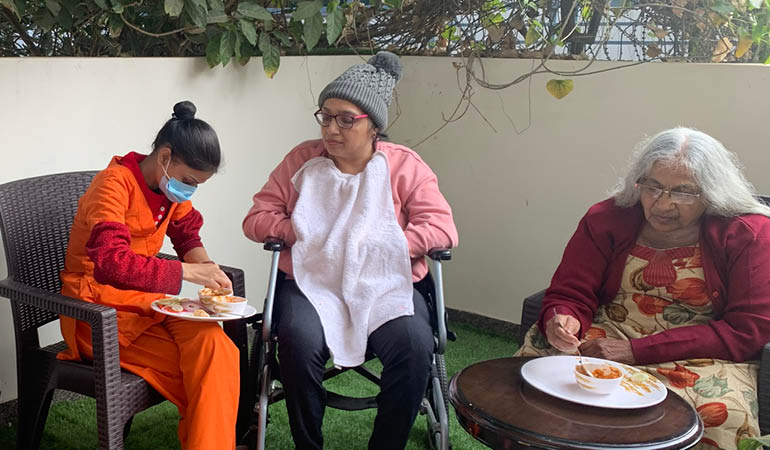 A professional caregiver assisting a senior woman with gentle mobility in a brightly lit hallway of an Assisted Living home for elderly in India.